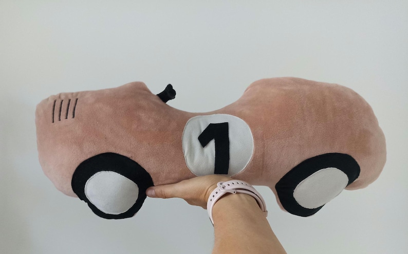 May include: A plush toy shaped like a vintage race car in a dusty pink color. The car features a white circular patch on the side with a black number one, black and white wheels, and four black stitched lines on the front hood area.