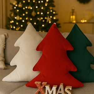Könnte beinhalten: Drei baumförmige Dekokissen in den Farben Creme, Rot und Dunkelgrün. Vor dem roten Kissen stehen vier einzelne Holzbuchstaben, die das Wort XMAS bilden, wobei das X mit weißen Punkten und das S mit gestreiften Linien versehen ist.