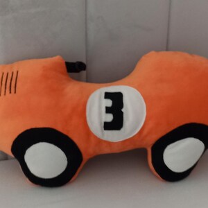 May include: A plush orange race car pillow featuring a white circular emblem on the side with the black number 3 printed in the center. The car design includes black and white wheels and four black vertical lines on the front hood area.