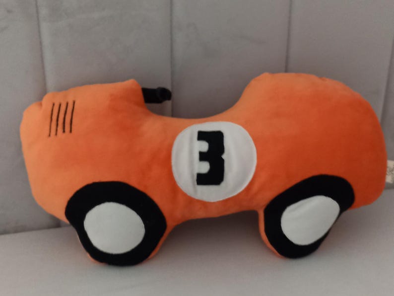 May include: A plush orange race car pillow featuring a white circular emblem on the side with the black number 3 printed in the center. The car design includes black and white wheels and four black vertical lines on the front hood area.