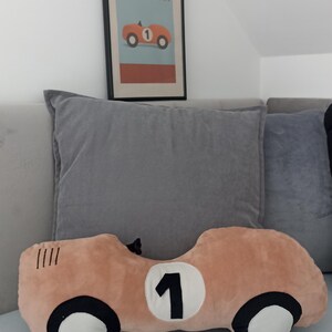 May include: A plush pillow shaped like a vintage race car in a light peach color with black and white wheels and a white circle containing the black number 1. Above it hangs a framed print featuring a race car and the text LITTLE CHAMPION.