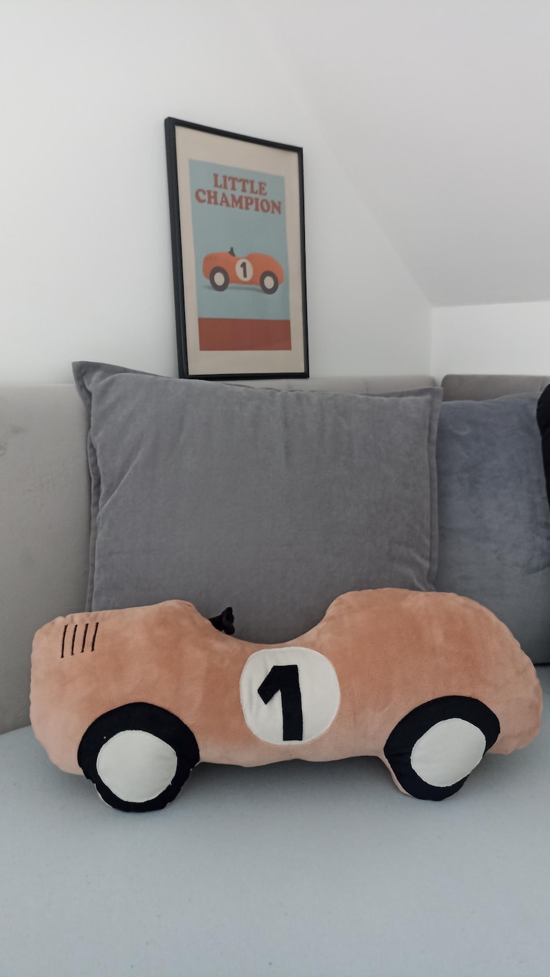 May include: A plush pillow shaped like a vintage race car in a light peach color with black and white wheels and a white circle containing the black number 1. Above it hangs a framed print featuring a race car and the text LITTLE CHAMPION.