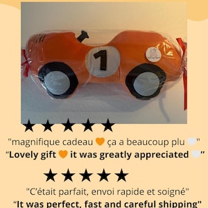 May include: An orange race car shaped plush pillow featuring a white circular patch with the black number 1, black and white wheels, and a small circular logo tag, all enclosed in clear plastic packaging with red ribbon ties on the sides.