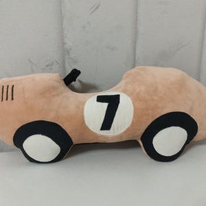 May include: A plush car-shaped pillow in a soft tan color. The design features black and white wheels, a black steering wheel detail, and a white circular patch on the side with the black number 7 printed in the center of the vehicle body.