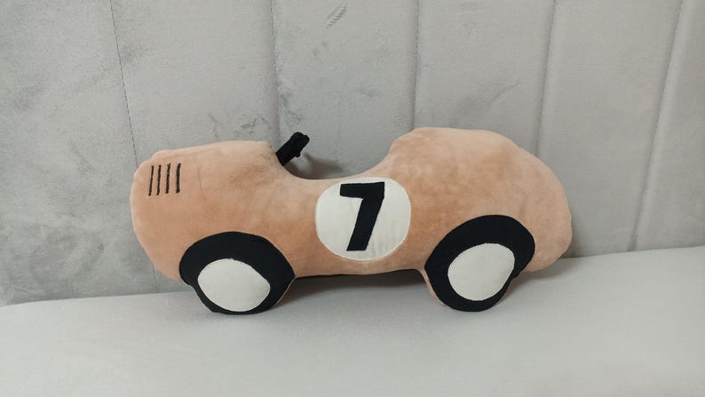 May include: A plush car-shaped pillow in a soft tan color. The design features black and white wheels, a black steering wheel detail, and a white circular patch on the side with the black number 7 printed in the center of the vehicle body.