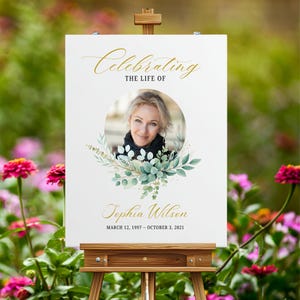 May include: A white sign on a wooden easel with the text "Celebrating The Life Of" in gold script. A photo of a woman is surrounded by a floral wreath. The name "Tephia Wilson" and dates are below the photo.