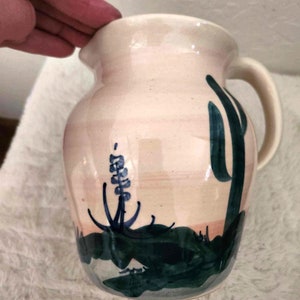 Könnte beinhalten: Ein weißer Keramik Krug mit einer grünen und blauen Wüstenlandschaft, die auf die Seite gemalt ist. Die Szene zeigt einen hohen Kaktus und eine kleinere Pflanze mit blauen Blüten.