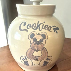 May include: A beige ceramic cookie jar with a lid. The jar is decorated with the word "Cookies" in dark blue script and a cartoon bear illustration. The bear is brown and tan with dark blue accents.