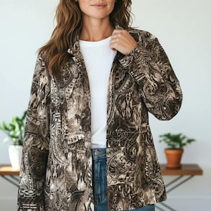 May include: A brown and beige paisley print blazer. The open-front jacket is worn over a white t-shirt and blue jeans. The blazer features a classic collar and long sleeves. The background includes a wooden table and potted plants.