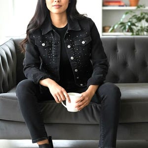 May include: A black denim jacket with rhinestone embellishments, paired with black jeans and black ankle boots. The person is holding a white mug while sitting on a gray sofa. The outfit is all black.