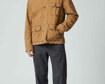 May include: A tan work jacket with four front pockets and button closures. The jacket is paired with dark gray pants and white sneakers. The model is wearing a black t-shirt underneath the jacket.