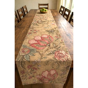 May include: A beige floral table runner with large pink and red flowers and green leaves. The runner is placed on a long, wooden table. The table is surrounded by wooden chairs. A bowl of green apples sits at the table's center.