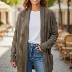 Könnte beinhalten: Eine Frau trägt einen langen, olivgrünen Cardigan mit Rippstruktur, ein weißes T-Shirt und blaue Jeans. Der Cardigan hat zwei Vordertaschen. Der Hintergrund zeigt eine verschwommene Außenkulisse mit Bäumen und Gebäuden.