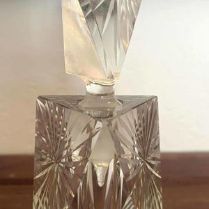 May include: A clear glass bottle with a geometric, faceted design. The bottle has a square base and a triangular stopper. The bottle is decorated with a starburst pattern.