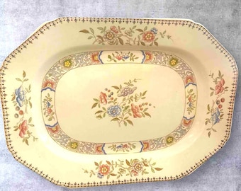 Puede incluir: Plato de servir octogonal de color crema con diseño floral y de volutas. El plato presenta un óvalo central con un ramo de flores en tonos rosa, azul y melocotón. El borde tiene un patrón repetitivo de pequeños puntos y motivos florales.