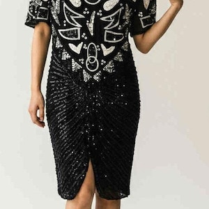 May include: A black sequined dress with a short-sleeved top adorned with an abstract white and silver design. The knee-length skirt has a front slit. The outfit is completed with black strappy heels.