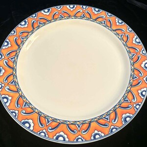 May include: A round, white ceramic plate with a decorative orange and blue border. The border features a repeating pattern of stylized floral and geometric shapes. The plate is suitable for serving food.