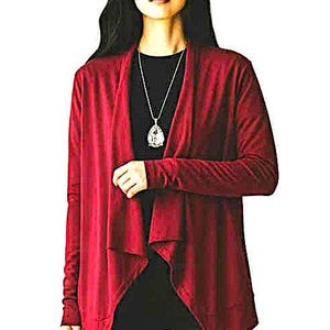 Puede incluir: Un cárdigan rojo oscuro con mangas largas. El diseño frontal abierto cae elegantemente. El cárdigan se usa sobre una blusa negra y pantalones negros. También es visible un collar plateado con un colgante.