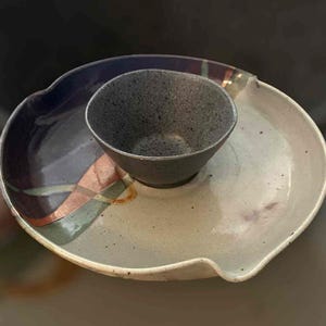 Puede incluir: Una fuente de cerámica hecha a mano con un cuenco adjunto. La fuente ovalada tiene una base beige claro y un borde azul oscuro. El cuenco es de color gris oscuro. La fuente tiene un patrón de ondas decorativas en tonos naranja, verde y morado.