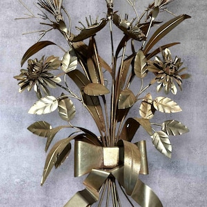 Può includere: Scultura murale floreale in metallo dorato con fiori stilizzati, foglie e un grande fiocco. La scultura ha un'estetica vintage ed è montata su una parete grigia. Il metallo ha un aspetto leggermente ossidato.