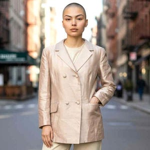 May include: A light beige double-breasted blazer with two rows of buttons and front pockets. The blazer is worn with matching beige trousers and a cream-coloured top. The outfit is worn outdoors.