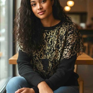 Op de afbeelding: Een zwarte trui met gouden bloemen kanten details op het bovenste gedeelte. De trui heeft lange mouwen en een ronde hals. De persoon op de foto draagt een blauwe spijkerbroek en zit op een houten stoel.