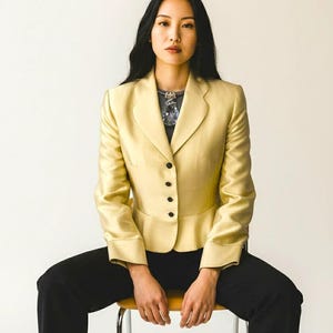 May include: A light yellow blazer with black buttons and long sleeves. The blazer is paired with black pants. The person is wearing a necklace with a large pendant. The person is sitting on a wooden chair with metal legs.