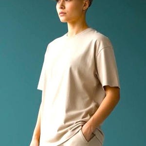 May include: A person wearing a beige t-shirt, matching pants, and a brown and black plaid newsboy cap. The outfit is a neutral color and the background is a teal color.