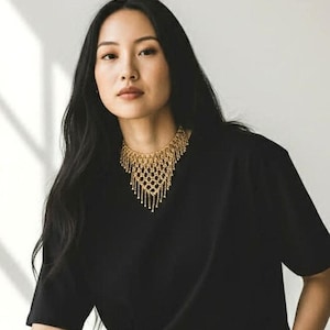 May include: A woman wearing a gold-colored statement necklace with a geometric design and dangling accents. She is wearing a black top. The background is a neutral color.