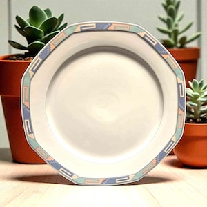 May include: White octagonal plate with a decorative border featuring a geometric pattern in pastel colors of blue, peach, and green. The plate is clean and appears unused, suitable for serving food.