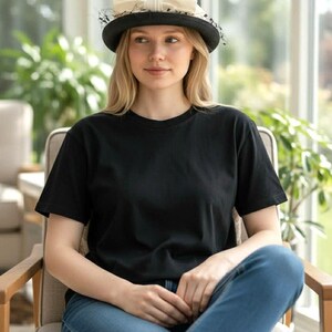 May include: A black hat with a cream-colored bow and netting detail. The hat is worn by a person wearing a black t-shirt and blue jeans. The person is sitting in a chair.