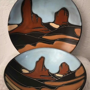 Könnte beinhalten: Zwei Keramikteller mit einem Wüstenlandschaft-Design. Der größere Teller hat einen blauen Himmel und der kleinere Teller einen hellblauen Himmel. Beide Teller sind in Braun, Schwarz und Weiß gehalten.