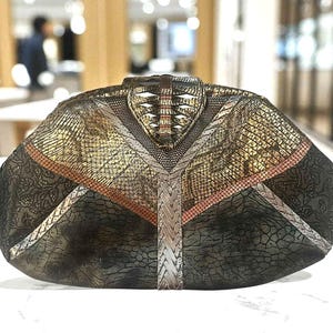 May include: A unique, textured clutch with a geometric design. The bag features a mix of metallic gold, silver, and bronze tones, with a reptile-like pattern. Accents include a woven silver strip and a patterned, dark green section. The clutch has a distinctive, angular shape.