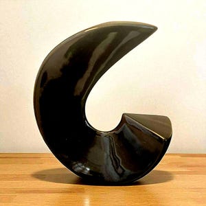 以下が含まれることがあります： 光沢のある黒いセラミック彫刻で、曲線的で抽象的なデザインです。彫刻は滑らかで反射する表面を持ち、木製の表面に置かれています。その形は、様式化された文字や波を連想させます。