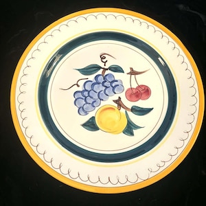May include: A round, decorative plate with a yellow rim and a white center. The center features a painted design of blue grapes, red cherries, and a yellow peach. A dark blue and green band encircles the fruit design.