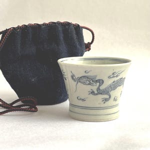 Könnte beinhalten: Eine weiße Keramiktasse mit blauem Drachendesign und einem dunkelblauen Beutel mit Kordelzug. Die Tasse hat einen ausgestellten Rand und einen zylindrischen Boden. Der Drache ist in traditioneller, stilisierter Weise dargestellt.