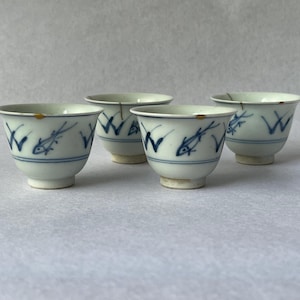 May include: Four small, white ceramic teacups with blue fish and wave designs. Each cup has a rounded base and a slightly flared rim. The cups are arranged on a white surface.