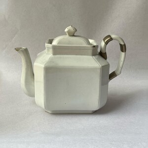 Könnte beinhalten: Eine weiße Keramik-Teekanne mit geometrischem Design. Die Teekanne hat einen Deckel mit rautenförmigem Knauf, einen gebogenen Ausguss und einen Griff mit goldenen Akzenten. Ein klassisches Design.