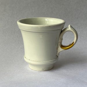 Könnte beinhalten: Cremefarbene Keramiktasse mit ausgestelltem Korpus und rundem Henkel. Der Henkel hat einen goldfarbenen Akzent. Die Tasse hat ein klassisches Design, geeignet für Kaffee oder Tee. Die Tasse ist etwa 10 cm hoch.