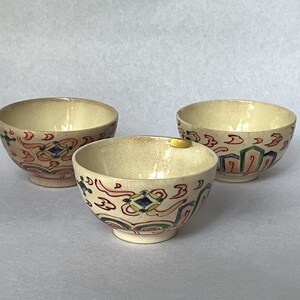 May include: Three small, off-white ceramic bowls with a decorative pattern of red, blue, and green shapes. The bowls have a rounded shape and a small footed base. The interior of the bowls is a light beige color.