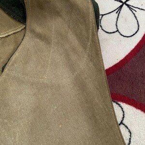 Peut inclure: Gros plan d'un gilet beige avec un col vert fonc&eacute;. Le gilet a une texture douce, semblable &agrave; du daim, et des d&eacute;tails de couture visibles. Le fond est un tissu &agrave; motifs avec des couleurs blanches, noires et bordeaux.