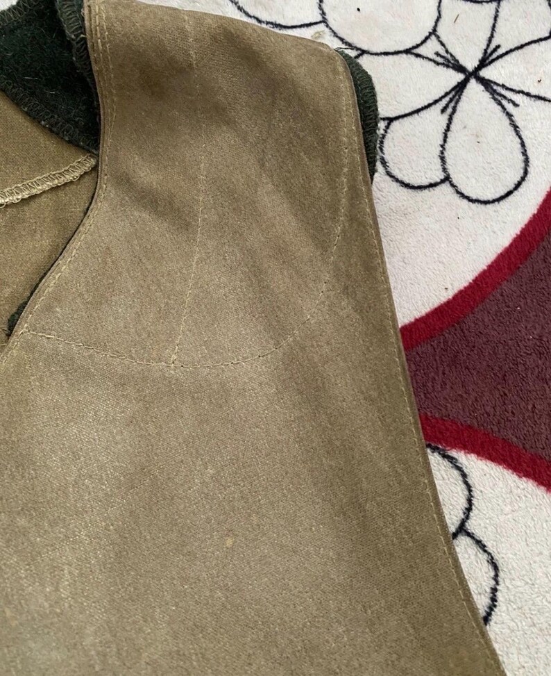 Peut inclure: Gros plan d'un gilet beige avec un col vert fonc&eacute;. Le gilet a une texture douce, semblable &agrave; du daim, et des d&eacute;tails de couture visibles. Le fond est un tissu &agrave; motifs avec des couleurs blanches, noires et bordeaux.