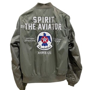 May include: An olive green bomber jacket featuring white embroidered text reading SPIRIT OF THE AVIATOR. The back displays a circular patch with a blue bird emblem, a red star, and the text 57th Fighter Weapons Wing, U.S. Air Force Nellis AFB, Thunderbirds, and AVIREX LTD.