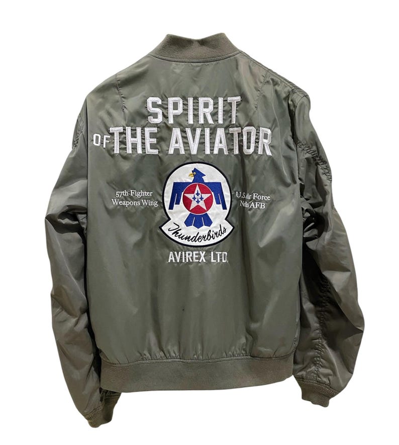 May include: An olive green bomber jacket featuring white embroidered text reading SPIRIT OF THE AVIATOR. The back displays a circular patch with a blue bird emblem, a red star, and the text 57th Fighter Weapons Wing, U.S. Air Force Nellis AFB, Thunderbirds, and AVIREX LTD.