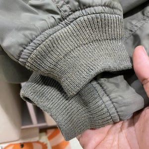 May include: An olive green jacket sleeve featuring a ribbed knit cuff. The sleeve is constructed from a smooth, solid-colored fabric with visible stitching along the seam where the cuff attaches to the main body of the garment.