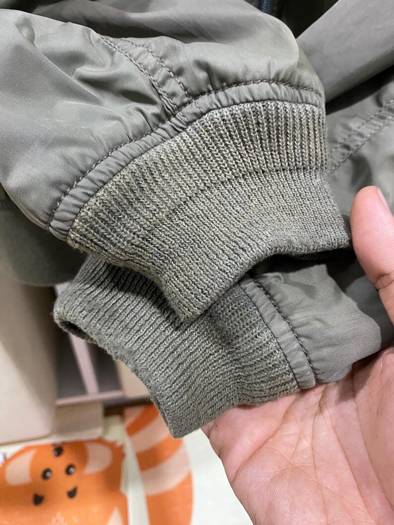 May include: An olive green jacket sleeve featuring a ribbed knit cuff. The sleeve is constructed from a smooth, solid-colored fabric with visible stitching along the seam where the cuff attaches to the main body of the garment.