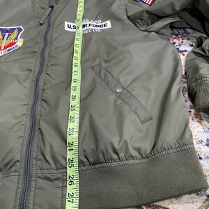 May include: An olive green bomber jacket with a front zipper, ribbed cuffs, and hem. It features patches including a U.S. flag on the shoulder and a U.S. Air Force emblem. A yellow measuring tape shows a length of approximately 29 inches along the front.