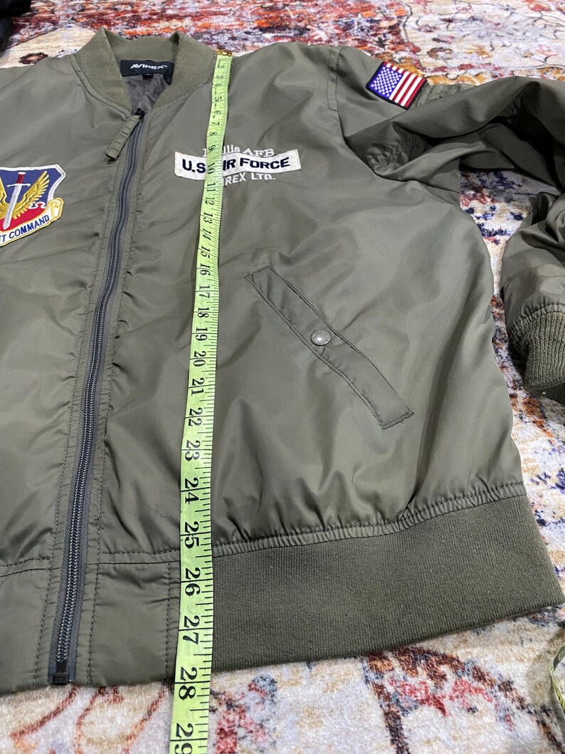May include: An olive green bomber jacket with a front zipper, ribbed cuffs, and hem. It features patches including a U.S. flag on the shoulder and a U.S. Air Force emblem. A yellow measuring tape shows a length of approximately 29 inches along the front.