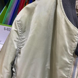 May include: Close-up of a light green bomber jacket. The jacket has a ribbed gray collar and a zipper closure. The fabric has a slight sheen, and the stitching is visible along the seams. The jacket has two pockets.