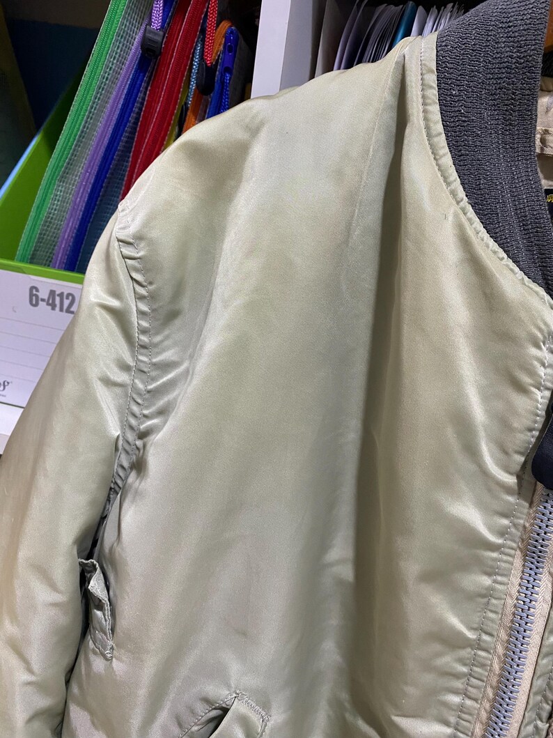 May include: Close-up of a light green bomber jacket. The jacket has a ribbed gray collar and a zipper closure. The fabric has a slight sheen, and the stitching is visible along the seams. The jacket has two pockets.